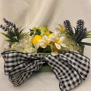 Puede incluir: Arreglo floral decorativo en un recipiente rectangular verde. El arreglo incluye flores blancas y amarillas, limones y lavanda. Un gran lazo a cuadros blanco y negro está sujeto al frente del recipiente.