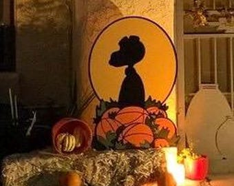 May include: A decorative wooden cutout featuring a black silhouette of a dog against a large yellow moon, positioned behind a cluster of orange pumpkins with green leaves. The display is set on a textured surface with additional pumpkins.