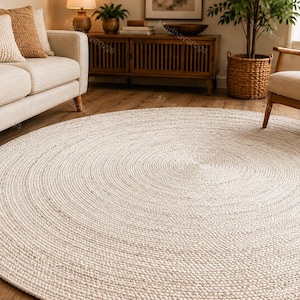 May include: A large, circular area rug featuring a braided, off-white texture. The rug is constructed from tightly coiled, woven fibers that create a consistent, concentric pattern across its entire surface, providing a neutral foundation.