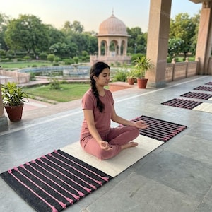 May include: A rectangular woven rug featuring a central cream-colored section flanked by two black ends decorated with vertical pink stripes and small pink tassels. The rug is laid flat on a stone floor, providing a surface for seated meditation.