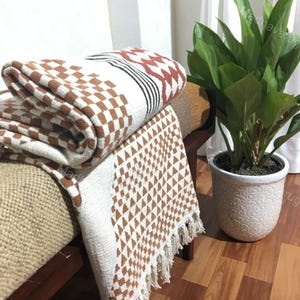 Puede incluir: Una manta tejida doblada que presenta una base blanca con patrones de cuadros marrones, rayas negras y un diseño geométrico triangular marrón a lo largo del borde, acabado con borlas de flecos color crema en el dobladillo inferior.