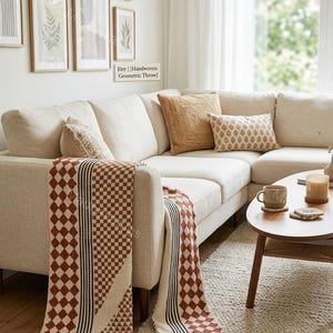 May include: A handwoven throw blanket featuring a geometric pattern of brown diamonds and black stripes on a cream background with fringe edges, draped over a beige sectional sofa with several decorative throw pillows and a wooden coffee table.