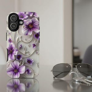 May include: A smartphone case featuring a white background with embossed-style purple flowers and silver swirling vine patterns. The case has a black camera lens cutout at the top left corner, showcasing a floral design with depth and texture.