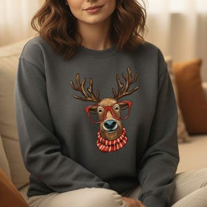 May include: A charcoal gray crewneck sweatshirt featuring a central graphic of a brown reindeer with large antlers, wearing bright red oversized glasses and a red and white striped scarf around its neck, set against a solid dark gray fabric background.