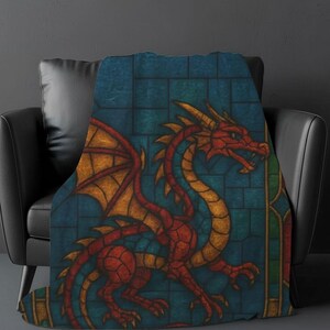 May include: A soft throw blanket featuring a vibrant stained glass style illustration of a dragon. The dragon is rendered in shades of red and gold against a deep blue grid background, with sharp claws, a long tail, and large wings displayed.