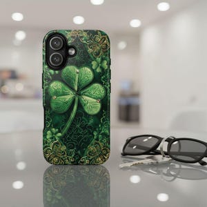 May include: A green phone case featuring a detailed shamrock design with gold accents. The case has a textured appearance and a dark green background. The shamrock is the focal point, with intricate detailing. The case is next to a pair of sunglasses and keys.