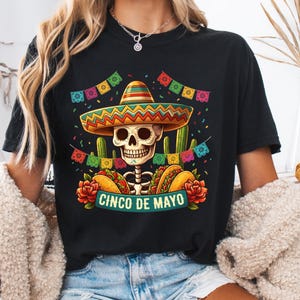 May include: A black t-shirt featuring a graphic of a skull wearing a colorful sombrero, surrounded by cacti, tacos, red roses, and festive bunting. The text CINCO DE MAYO is printed in a teal banner at the bottom of the graphic design.