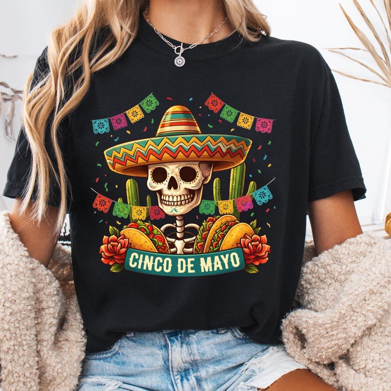 May include: A black t-shirt featuring a graphic of a skull wearing a colorful sombrero, surrounded by cacti, tacos, red roses, and festive bunting. The text CINCO DE MAYO is printed in a teal banner at the bottom of the graphic design.