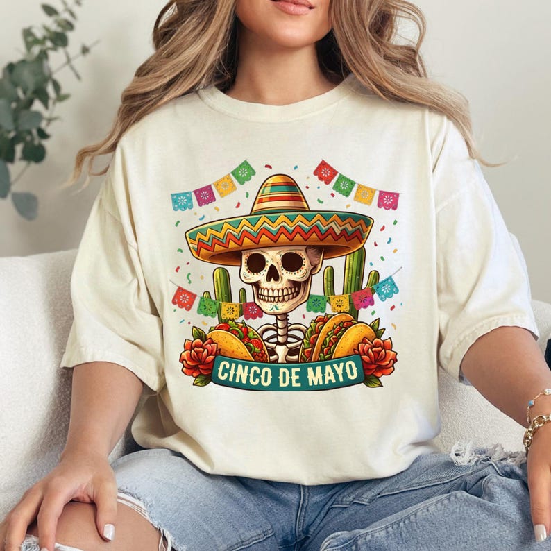 May include: A cream-colored t-shirt featuring a graphic of a skeleton wearing a colorful sombrero, surrounded by cacti, tacos, red roses, and festive papel picado banners. The text "CINCO DE MAYO" is printed in a teal banner at the bottom of the design.