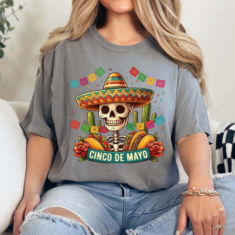 May include: A grey t-shirt featuring a graphic of a skeleton wearing a colorful sombrero, surrounded by cacti, tacos, red flowers, and festive bunting. The text "CINCO DE MAYO" is printed in a teal banner at the bottom of the graphic design.