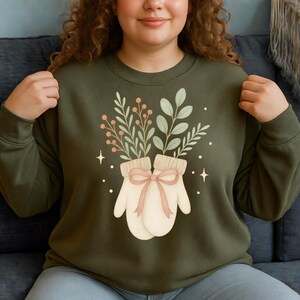 May include: Olive green sweatshirt featuring a graphic of two cream-colored mittens tied with a pink bow, filled with green leaves and red berries. The design includes small star accents. A cozy, nature-themed apparel item.