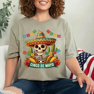 May include: A light olive green t-shirt featuring a graphic of a skeleton wearing a colorful sombrero, surrounded by cacti, tacos, red roses, and festive bunting. The text CINCO DE MAYO is printed in a banner across the bottom of the design.