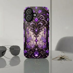 May include: A phone case with a stained-glass design in shades of purple, black, and white. The case features ornate, jewel-like embellishments and a camera cutout. Sunglasses and keys are visible in the background.