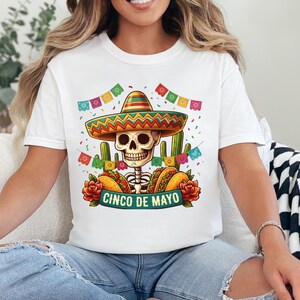 May include: A white t-shirt featuring a graphic of a skeleton wearing a colorful sombrero, surrounded by cacti, tacos, red flowers, and festive bunting. The text CINCO DE MAYO is printed on a teal banner at the bottom of the central illustration.