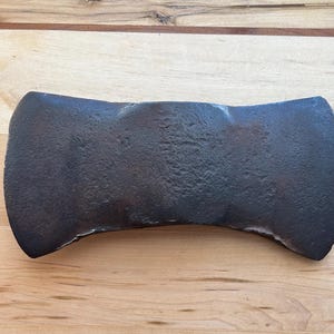 May include: A weathered, double-bit axe head, made of metal, with a dark finish. The axe head has a curved shape and two cutting edges. It is displayed on a light brown wooden surface, showing signs of age and use.