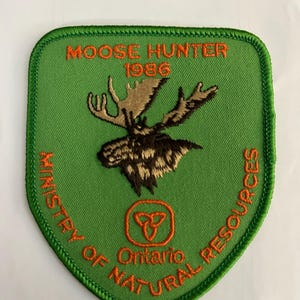 May include: Green patch with orange stitching featuring a moose head, the text "Moose Hunter 1986", and the logo for the Ministry of Natural Resources Ontario.