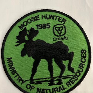 Peut inclure: Un écusson circulaire vert et noir avec le texte "Moose Hunter 1985 Ministry of Natural Resources" et une silhouette noire d'un élan. L'écusson comporte également un petit cercle blanc avec une feuille d'érable noire à l'intérieur et le mot "Ontario" en dessous.