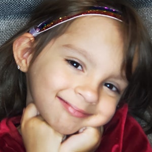 Puede incluir: Una niña pequeña con cabello largo y castaño lleva una camisa roja y una diadema brillante. Está sonriendo y mirando a la cámara.