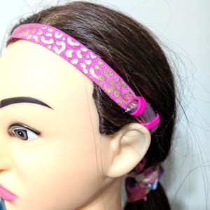 May include: Pink headband with a gold leopard print design. The headband is made of elastic material and is designed to be worn around the head.