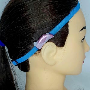 May include: A blue headband with a small pink hearing aid attached to the side.