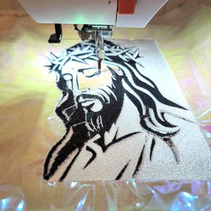 May include: An embroidery machine in operation, stitching a black and white image of Jesus wearing a crown of thorns onto a light-coloured fabric. The machine's needle is positioned over the design, with a close-up view of the detailed embroidery.