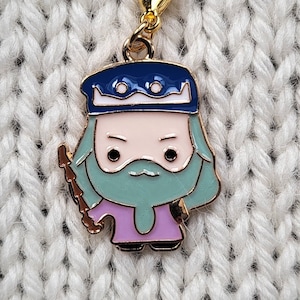 May include: A gold-colored charm featuring a cartoon character with a blue crown, a white beard, and a purple robe. The charm is on a white knitted background.
