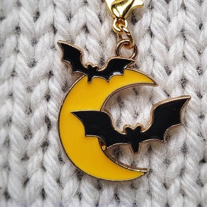 May include: A gold-colored charm featuring a yellow crescent moon with two black bats flying around it. The charm is attached to a gold-colored chain.