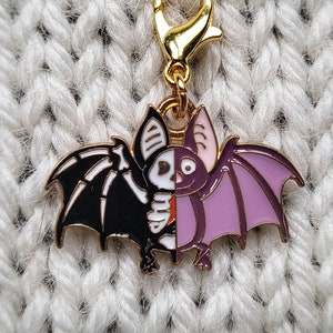 May include: A gold-tone metal charm featuring a black and white bat with a skeleton design on one side and a purple bat with a pink wing on the other side.