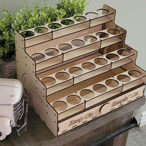 Puede incluir: Un organizador de madera sin terminar de varios niveles con múltiples aberturas circulares para almacenamiento. El organizador tiene un diseño escalonado y las palabras "Keep On" y "Keepin' On" están grabadas en el frente. Diseñado para objetos pequeños.