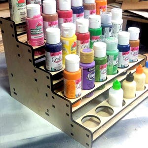 May include: A wooden paint bottle organiser with five shelves. The organiser is filled with various colours of acrylic paint bottles. The organiser is made of light brown wood and has a black trim.