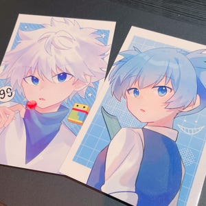 May include: Two anime-style art prints. One features a character with white hair, holding a lollipop, the other with blue hair, holding a knife. Both are set against a light blue grid background with a cartoonish style.