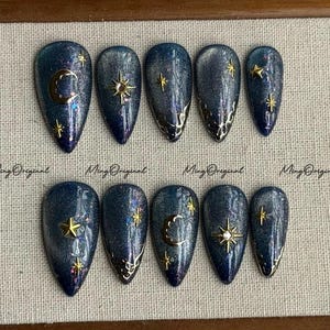 May include: A set of ten artificial nails with a dark blue glitter finish. Each nail features gold celestial designs, including stars and crescent moons. The text "Ming Original" is printed on the surface.