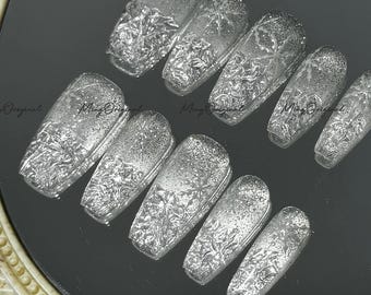 May include: A set of ten silver-colored, glittery, and textured artificial nails. The nails are arranged on a reflective surface, with a decorative floral pattern. The nails are in various sizes and shapes.