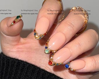 Könnte beinhalten: Eine Hand, die lange, mandelförmige Kunstnägel mit nackter Basis und juwelenbesetzten Goldspitzen zeigt. Die Nägel sind mit bunten Strasssteinen verziert. Die Hand trägt außerdem zwei goldene Ringe, einen mit einem grünen Stein und einen mit klaren Steinen.