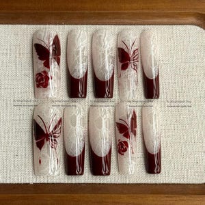 May include: A set of ten artificial nails with a white lace design, burgundy tips, and burgundy butterfly and rose accents. The nails are arranged on a light brown surface.