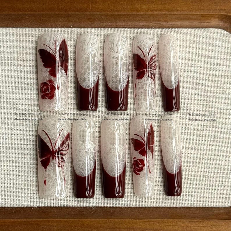 Pu&ograve; includere: Un set di dieci unghie finte con un design in pizzo bianco, punte bordeaux e motivi a farfalla e rosa bordeaux. Le unghie sono disposte su una superficie marrone chiaro.