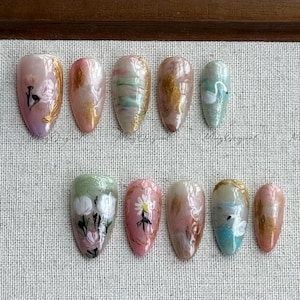 May include: A collection of ten artificial nails with various designs. The nails feature floral patterns, gold accents, and abstract swirls in shades of pink, green, and gold. Some nails have swan illustrations.