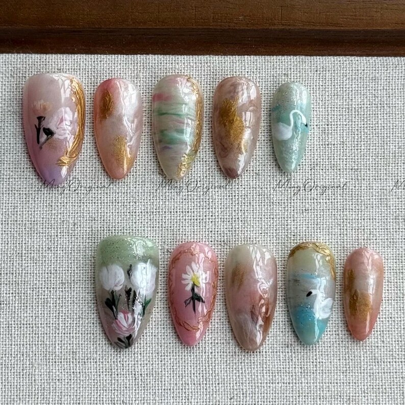 May include: A collection of ten artificial nails with various designs. The nails feature floral patterns, gold accents, and abstract swirls in shades of pink, green, and gold. Some nails have swan illustrations.