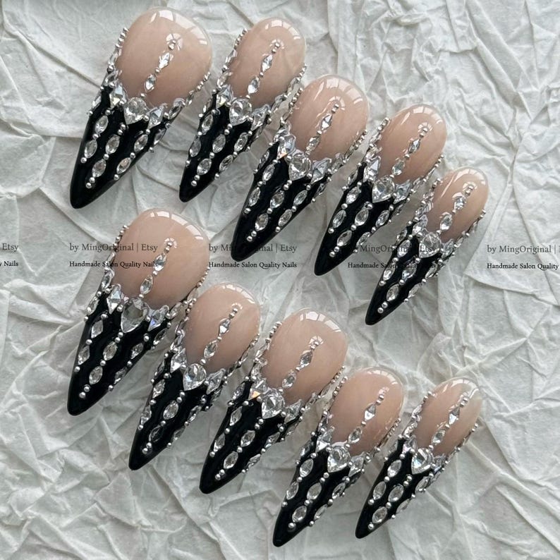 May include: A set of ten stiletto-shaped press-on nails featuring a nude base and a black tip. Each nail is decorated with clear, faceted rhinestones arranged in vertical lines and clusters, creating a sparkling, ornate design across the surface.
