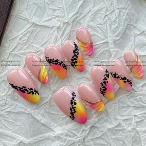May include: A set of ten almond-shaped press-on nails featuring a nude base with a gradient of yellow to pink at the tips. Some nails display a black leopard print pattern, while others feature raised gold-colored wavy lines across the surface.