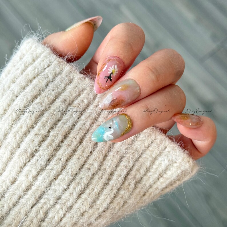 May include: Close-up of manicured nails with various designs. One nail features a white daisy with a black accent, another has a swan design, and others have abstract art. The nails are adorned with gold accents and set against a beige sweater.