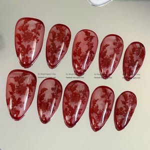 May include: A set of ten almond-shaped press-on nails in a translucent deep red finish, each featuring a detailed dark red floral lace pattern. The nails are arranged in two rows, displaying varying sizes with a glossy, smooth surface texture.