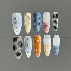 Pode incluir: Uma coleção de dez unhas artificiais com vários designs. As unhas apresentam cores como azul claro, bege, laranja e branco, com detalhes como tachas prateadas, bolinhas e padrões marmorizados. Algumas unhas têm um design texturizado 3D.