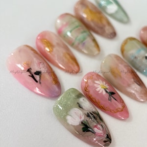 May include: A collection of painted artificial nails in various designs. The nails feature a range of colors, including pink, green, and gold, with floral and abstract patterns. The text "Ming Original" is visible on some nails.