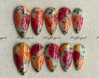 May include: Set of ten artificial nails with a floral design. The nails are almond-shaped with a clear base, featuring orange, pink, and white flowers. Each nail has a gold trim and the text "Ming Original".