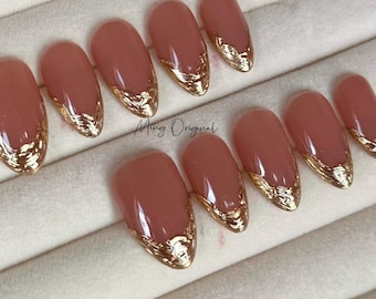 May include: Set of ten false nails with a nude base and gold accents. The nails are oval shaped and have a gold design on the free edge.
