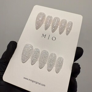 K&ouml;nnte beinhalten: Ein Set wei&szlig;er Glitzer-Press-On-N&auml;gel in verschiedenen Gr&ouml;&szlig;en. Die N&auml;gel werden auf einer wei&szlig;en Karte mit dem Text "M | O" und "www.mingoriginal.com" pr&auml;sentiert. Die N&auml;gel haben ein gl&auml;nzendes Finish und sind mit einer Spitze geformt.