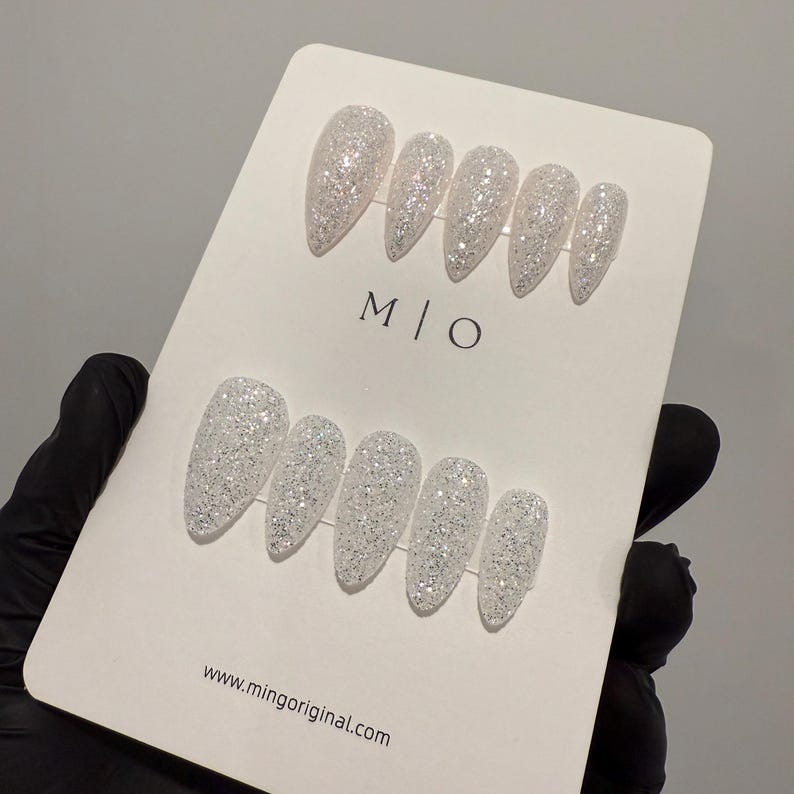 K&ouml;nnte beinhalten: Ein Set wei&szlig;er Glitzer-Press-On-N&auml;gel in verschiedenen Gr&ouml;&szlig;en. Die N&auml;gel werden auf einer wei&szlig;en Karte mit dem Text "M | O" und "www.mingoriginal.com" pr&auml;sentiert. Die N&auml;gel haben ein gl&auml;nzendes Finish und sind mit einer Spitze geformt.