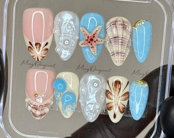 Puede incluir: Un conjunto de diez uñas postizas con temática de playa. Las uñas están decoradas con conchas marinas, estrellas de mar y burbujas. Las uñas están en una caja de plástico transparente con las palabras "Ming Original" impresas en la caja.
