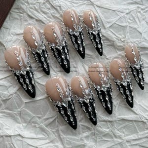 May include: A set of ten stiletto-shaped press-on nails featuring a nude base transitioning into a black tip. The nails are embellished with clear, faceted rhinestones arranged in a vertical pattern that extends from the nude area down to the black tips.